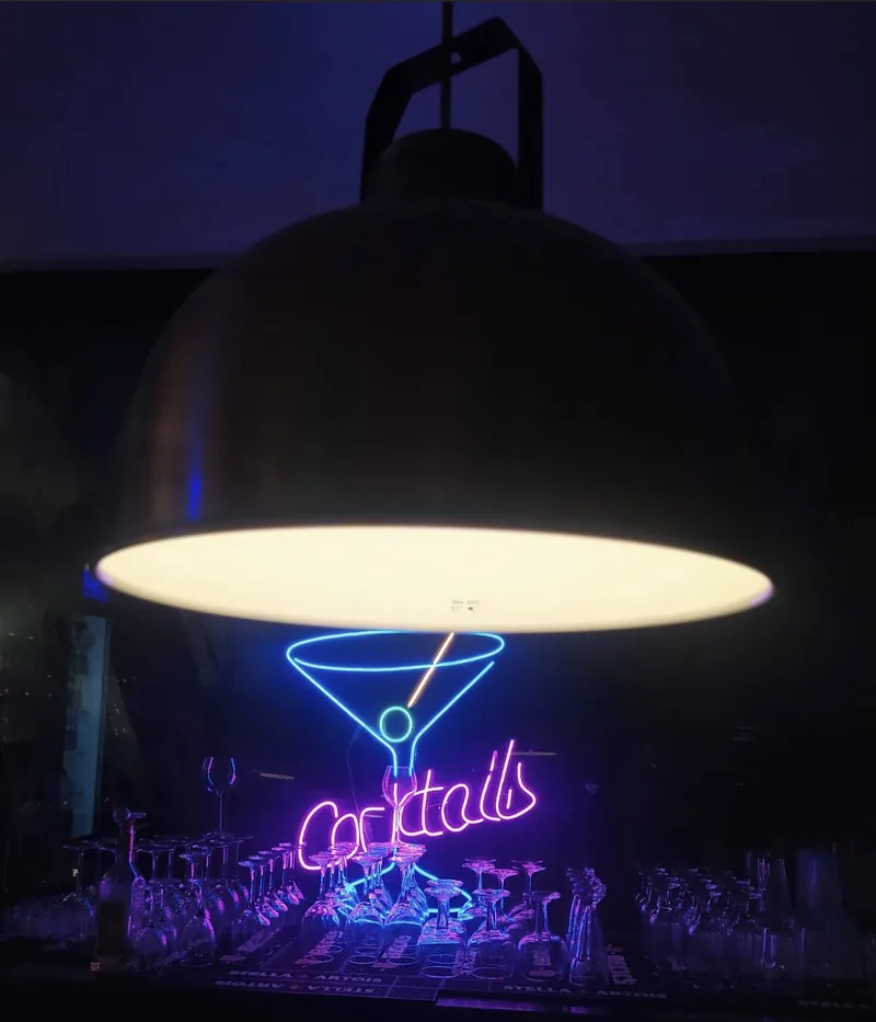 A neon “Cocktails” sign under a large hanging lamp, in the dark.